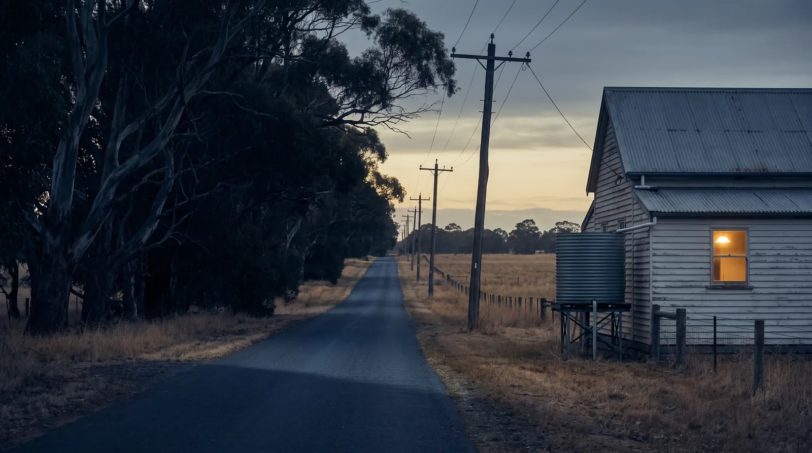 A country road with timber power poles and a farmhouse with one window lit