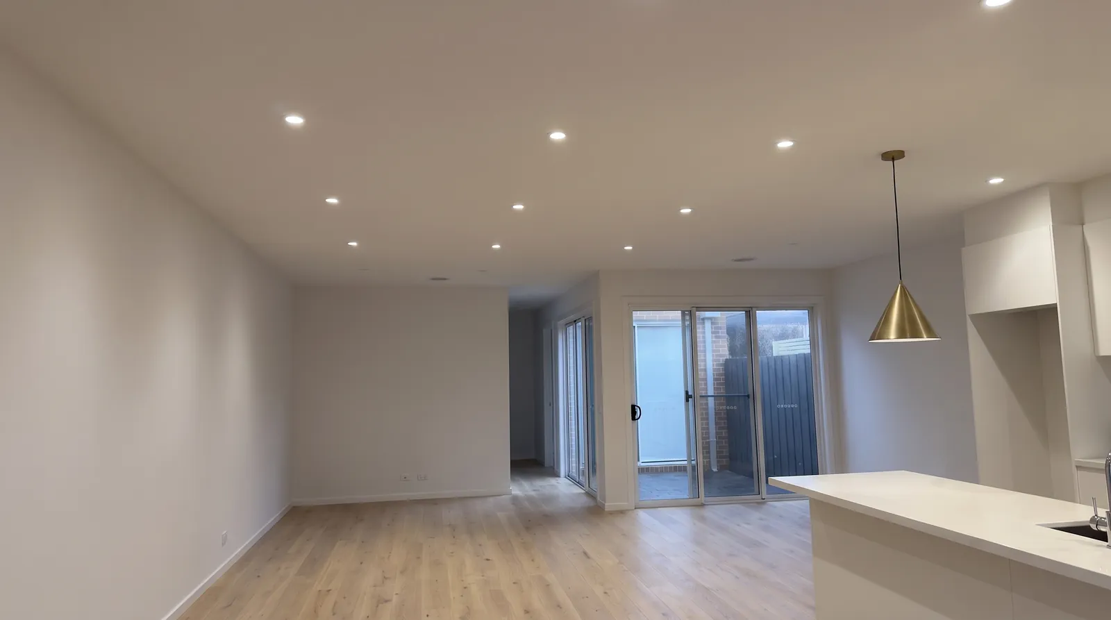 An open-plan living room with ceiling downlights on and a brass pendant over the bench
